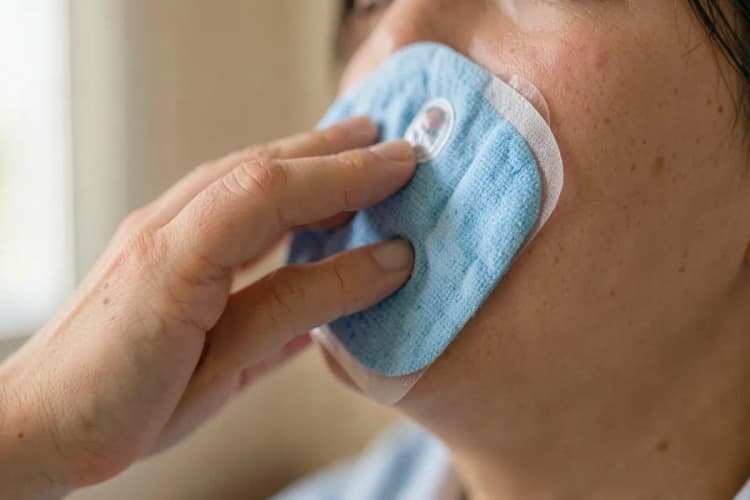 Pessoa aplicando cuidadosamente uma compressa fria na bochecha, na área de um implante dentário.